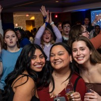 Three girls take a photo together while people behind them have their hands raised.